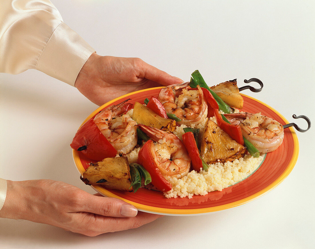 Hände halten Teller mit Shrimps-Ananas-Spiessen auf Reis