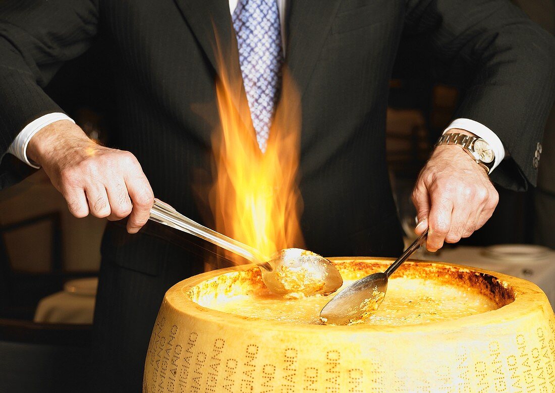 Hands Stirring a Flaming Wheel of Parmesan Cheese