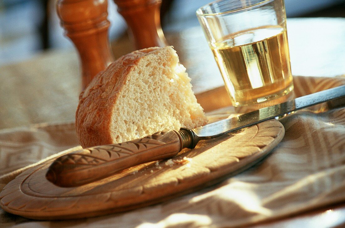 Stück Weißbrot auf Holzteller mit Messer, Glas Weißwein