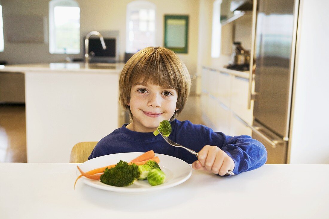 A Little Boy Holding Broccoli on a Fork in Front of a Plate of Mixed Vegetables