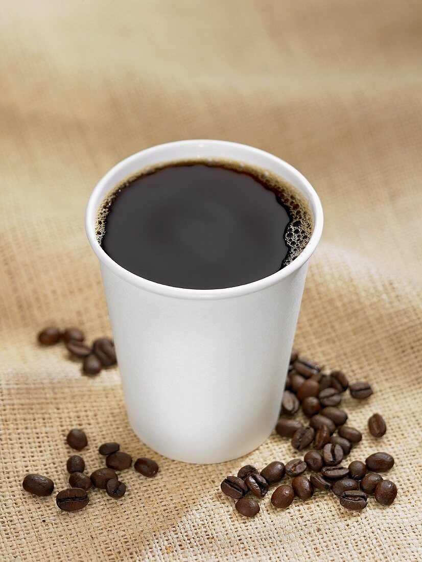 Schwarzer Kaffee im Plastikbecher, umgeben von Kaffeebohnen