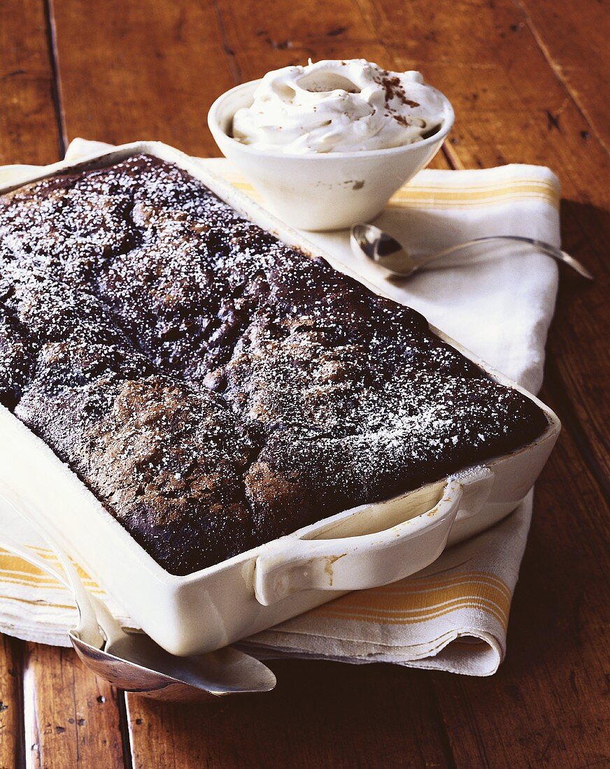 Chocolate Pudding Cake Topped with Powdered Sugar in Baking Dish; Bowl of Whipped Cream