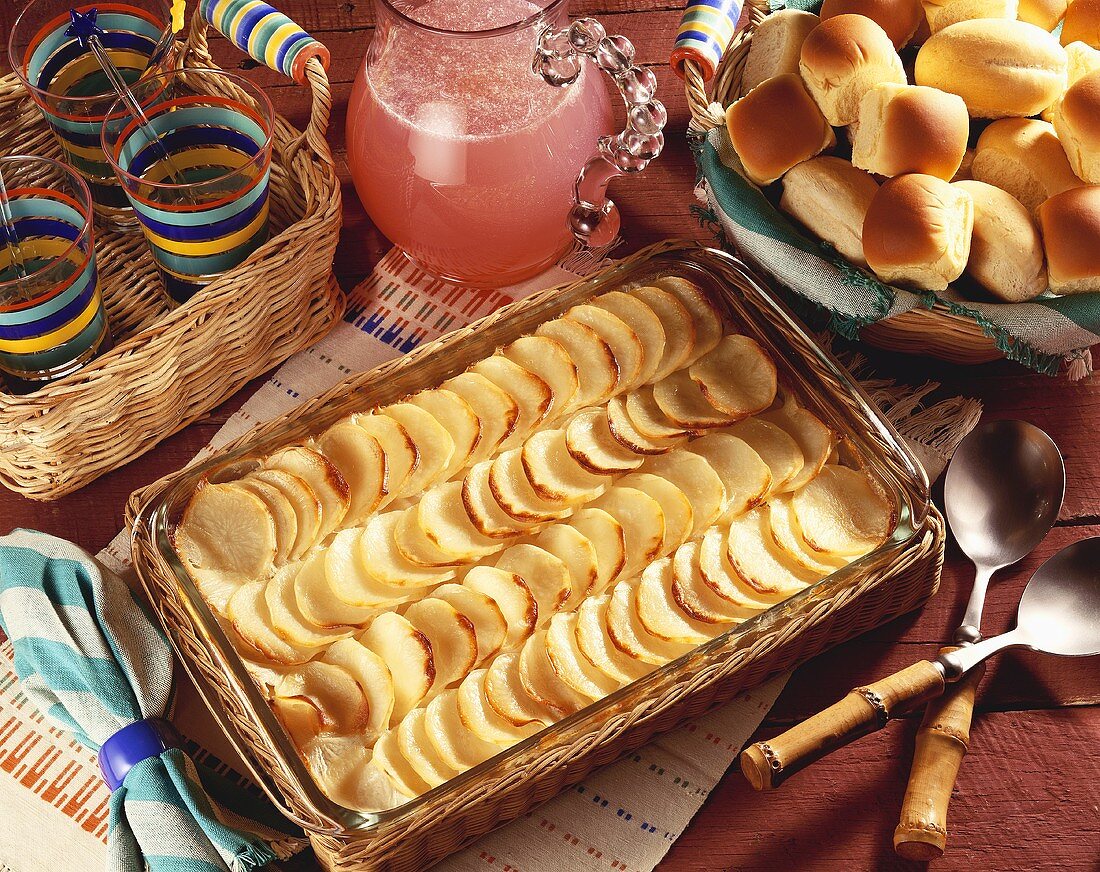 Scalloped Potatoes in Baking Dish on Picnic Table with Basket of Rolls and Pitcher of Pink Lemonade