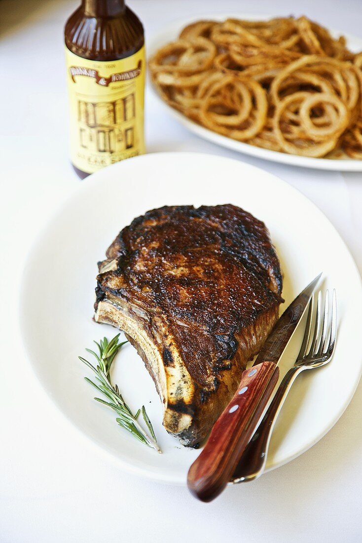 Rib Eye Steak on a White Plate; Rosemary Sprig; Knife and Fork; Onion Rings