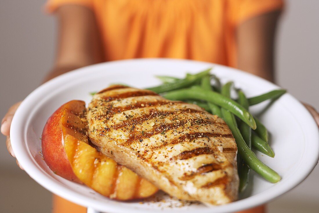 A girl holding a plate of grilled chicken, grilled peaches and beans