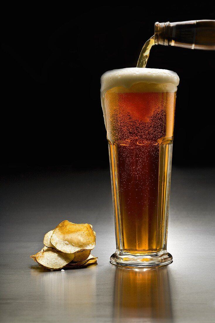 Pouring Amber Beer from a Bottle into a Tall Glass; Chips