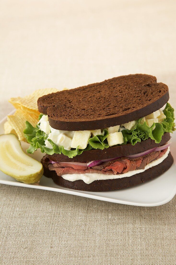 Pumpernickel-Clubsandwich mit Roastbeef; dazu Essiggurke und Chips