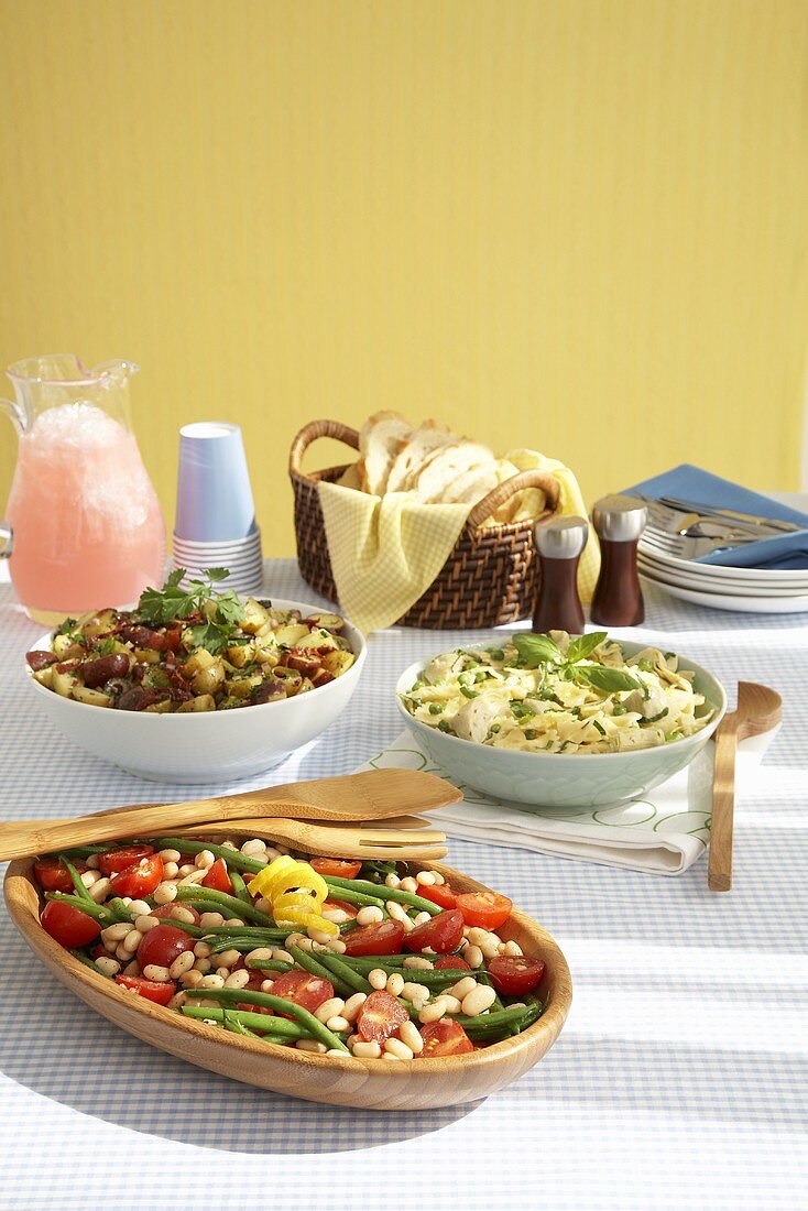 Three Salads in Serving Bowls on a Summer Table; Pitcher of Pink Lemonade; Basket of Bread