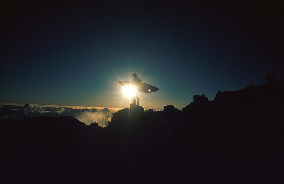 Silhouette eines Surfers mit Surfbrett auf Berggipfel im Gegenlicht eines Sonnenuntergangs
