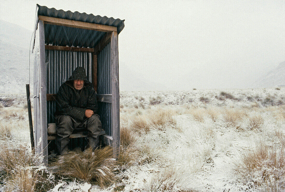 blocked for illustrated books in Germany, Austria, Switzerland: Musterer sitting on an outdoor toilet, Winter, High Country, South Island, New Zealand