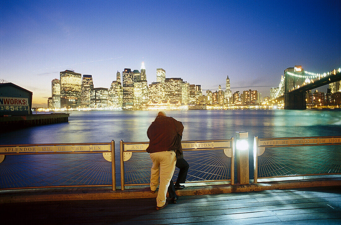Ein Paar auf der Brooklyn Bridge betrachtet die beleuchtete Skyline, Manhattan, New York, USA, Amerika