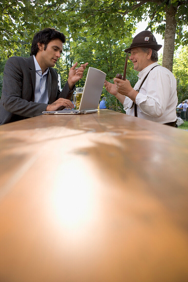 Old Bavarian man and young businessman with laptop in beer garden, Lake Starnberg, Bavaria, Germany