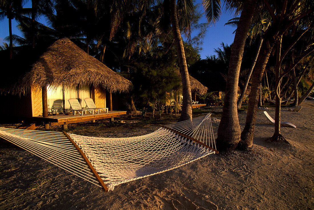 Hängematte zwischen Palmen vor Strandhütten in der Abendsonne, Manihi Pearl Beach Resort, Manihi Tuamotu, Französisch Polynesien