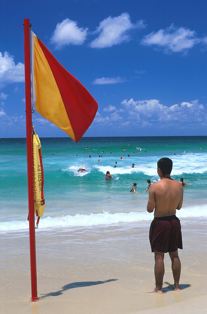 Ein Mann steht neben einer Fahne am Strand und beobachtet die Badenden, Bondi Beach, Sydney, New South Wales, Australien