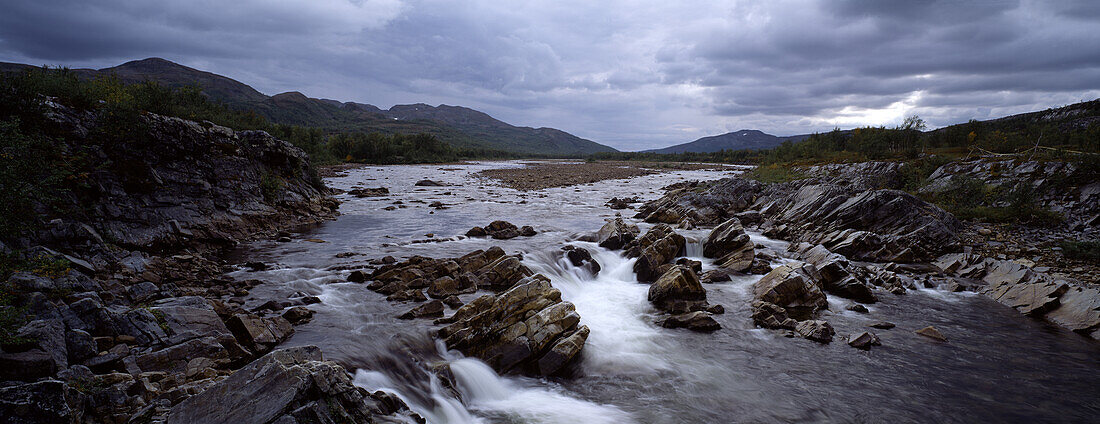 Ein Fluss und felsiges Ufer unter dunklen Wolken, Lappland, Schweden