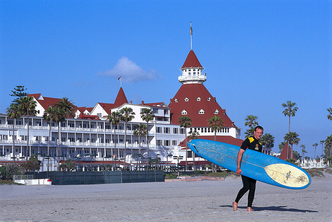 Surfer walking along the beach with surfboard, Hotel del Coronado, California USA