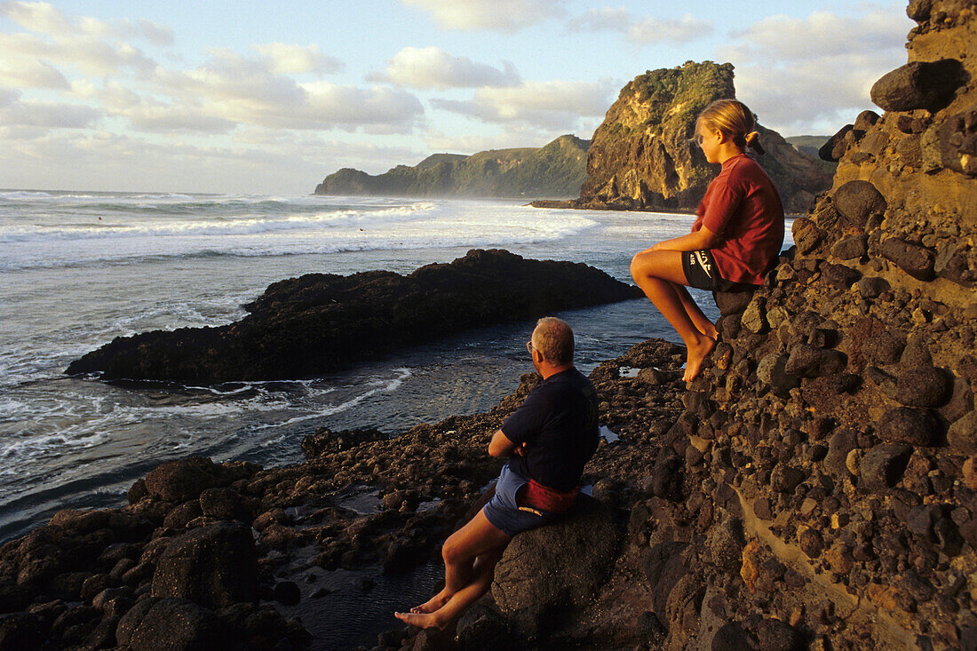 Tochter und Vater warten auf Sonnenuntergang, Strand Piha, Lion Rock, bekanntester Strand zum Surfen, West Küste in der nähe von Auckland [-], Nordinsel, Neuseeland