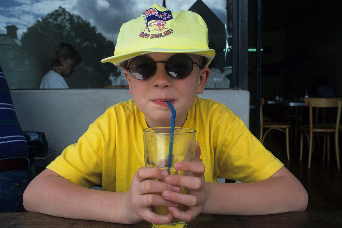 Portrait, young boy in yellow in cafe, Junge im Sonnenbrille und gelb trinkt Saft, boy with New Zealand cap drinking through straw, sunglasses