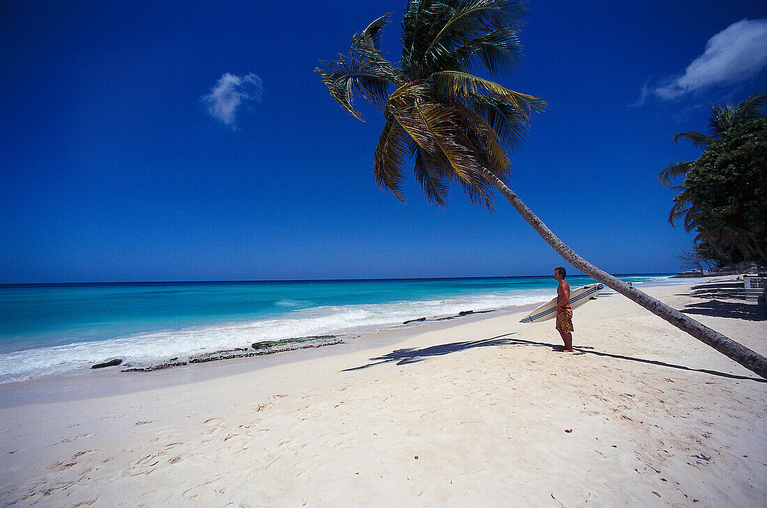 Mann mit Surfbrett steht am Palmenstrand, Barbados, Karibik, Amerika