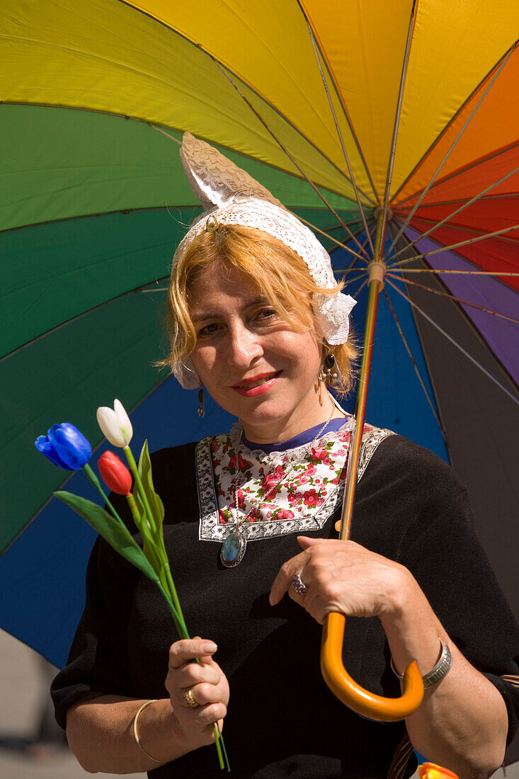 Holländische Frau in Tracht mit Schirm verkauft Tulpen auf Damm, Amsterdam, Niederlande