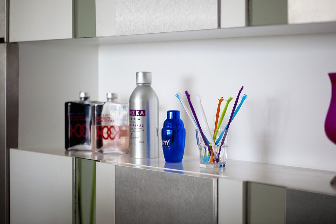 Flagons, bottles and a glass of coloured sticks on a shelf above a built-in cupboard