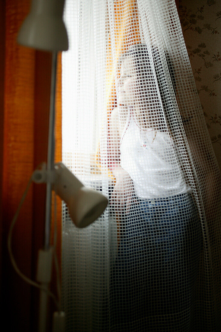 Young girl by curtain looking out window, front view