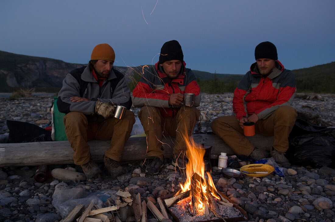 Drei maenner in der blauen stunde hinter einem lagerfeuer am Nahanni Fluss, Kanada