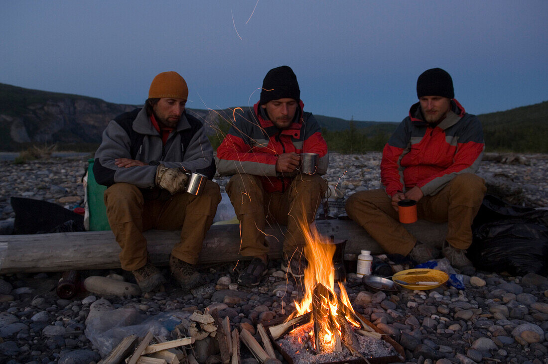 Three men sitting around a campfire at dusk, South Nahanni River, Canada