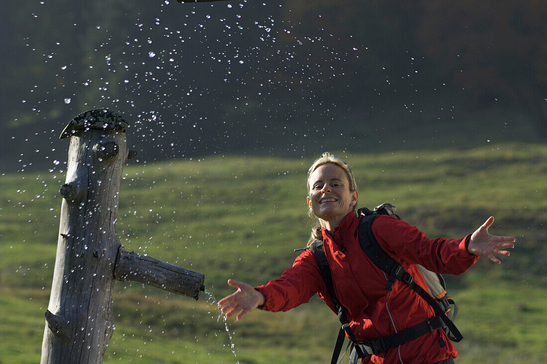 Junge Wandererin spritzt mit Wasser bei Brunnen auf Almwiese, Salzburger Land, Österreich