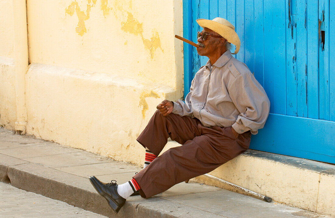 Blind old man sitting in front of a blue gate having a cigar, Havana, Kuba