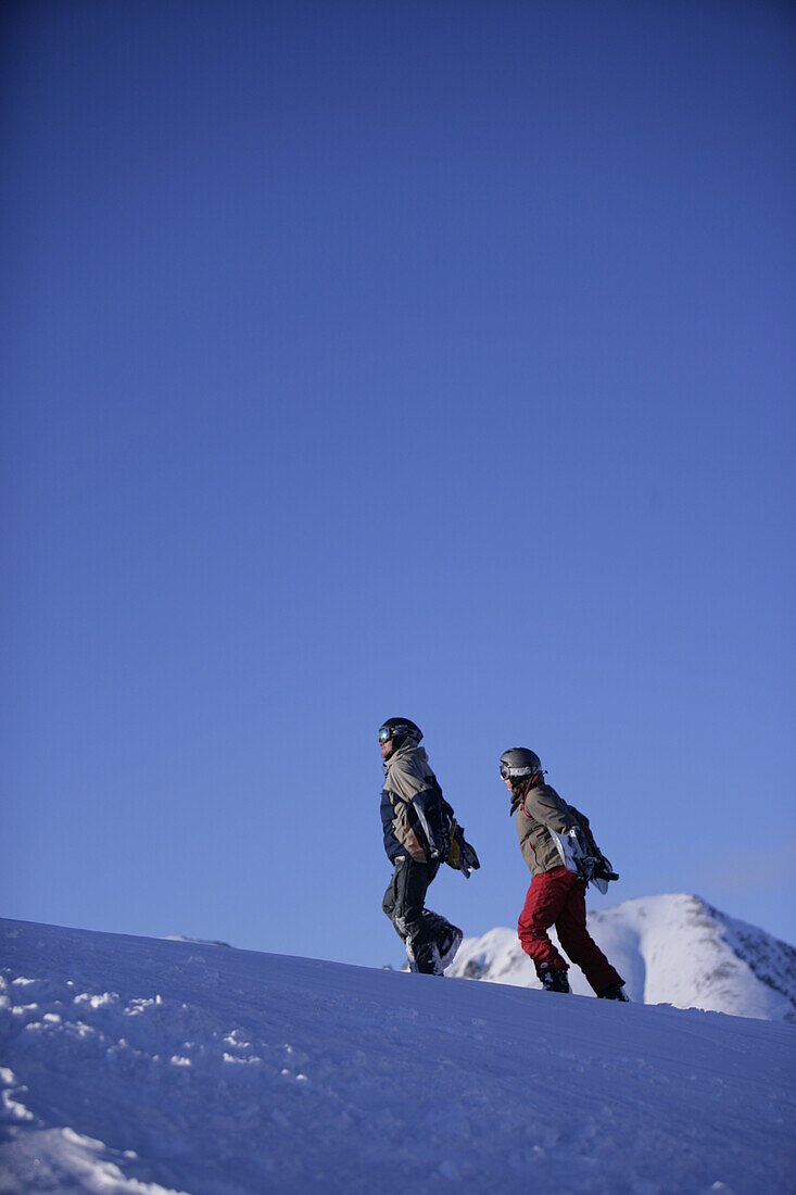 Two persons walking up a snowcapped mountain while carrying a snowboard, Kuehtai, Tyrol, Austria