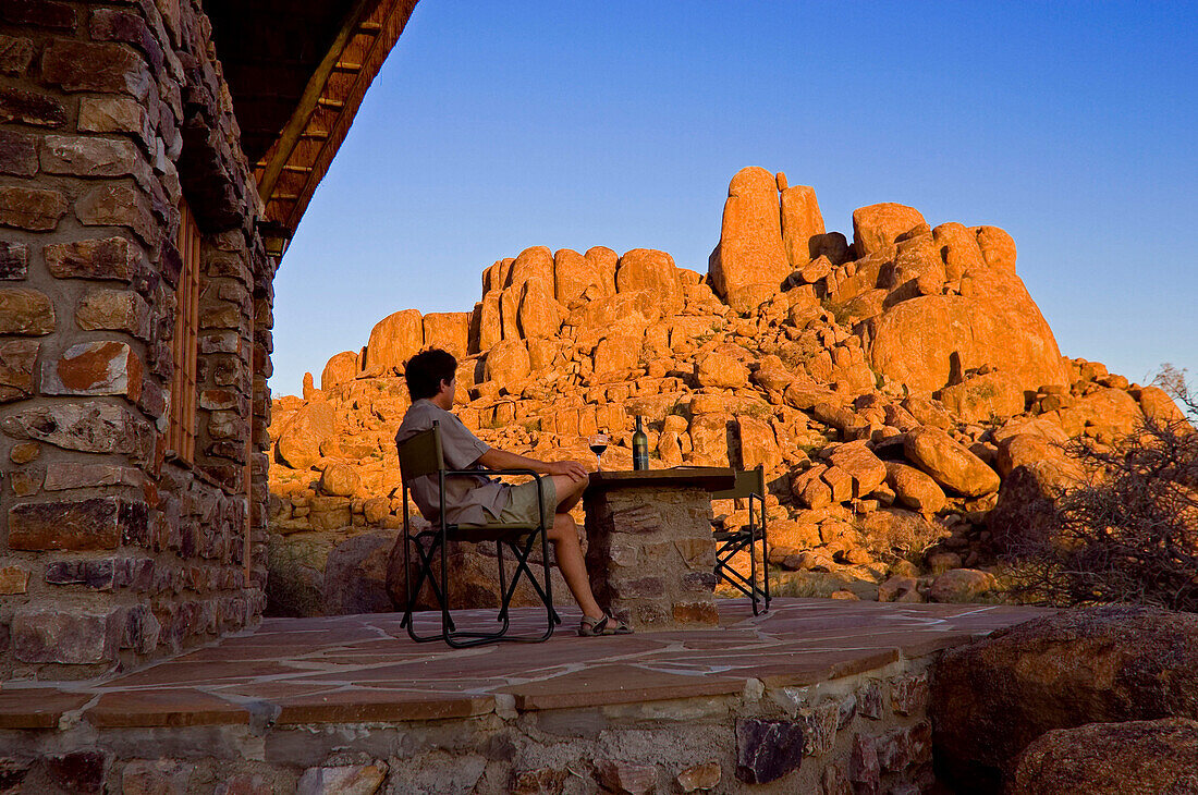 Junger Mann sitzt vor einem Bungalow und betrachtet die von der Abendsonne beschienenen Felsen. Canon Lodge,Gondwana Canon Park. Fish River Canyon, Südliches Namibia, Afrika. MR.