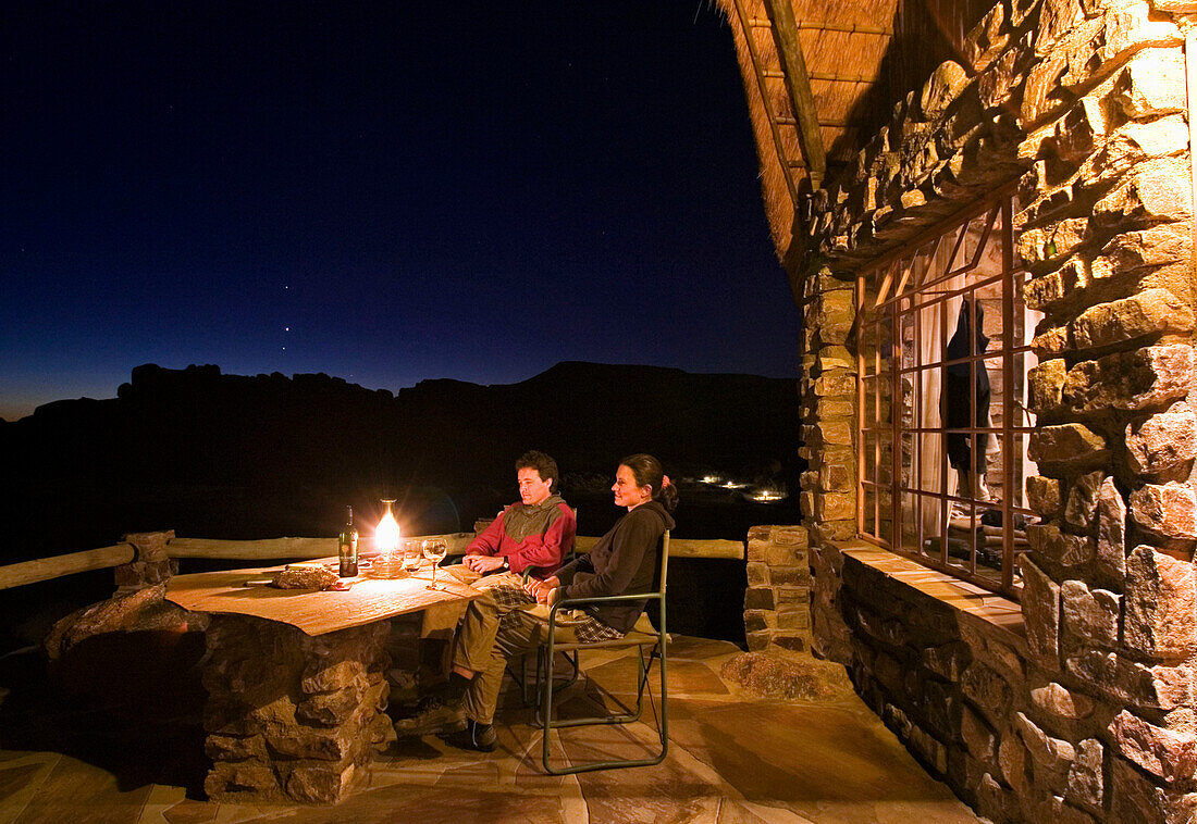 Ein junges Paar sitzt in der Nacht vor einem Bungalow. Auf dem Tisch eine Petrol Lampe. Canon Lodge, Gondwana Canon Park. Fish River Canyon, Südliches Namibia, Afrika, MR