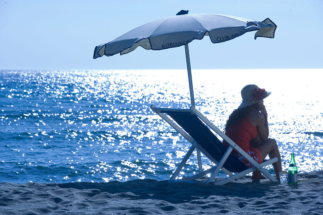 Woman sitting in deck chair on beach and drinking, Apulia, Italy