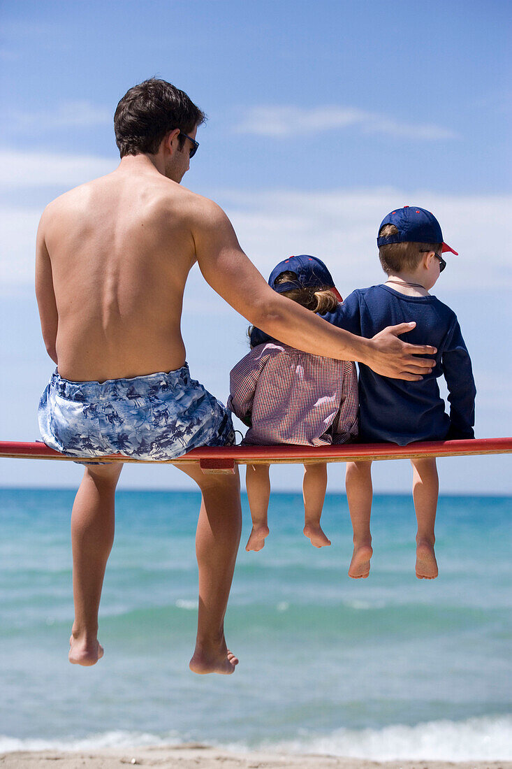 Junger Mann und zwei Kinder sitzen auf einer Bank am Strand, Apulien, Italien