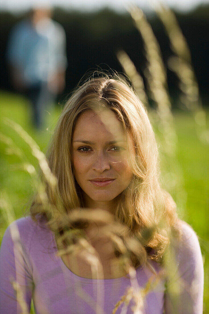 Young woman sitting on meadow, unrecognizable person in background