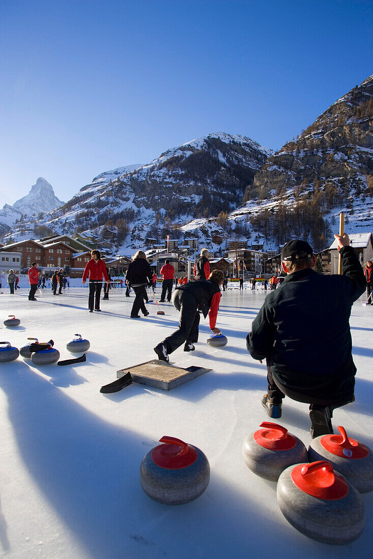 People curling on a rink, Matterhorn in background, Zermatt, Valais, Switzerland (Curling: A rink game where round stones are propelled by hand on ice towards a tee (target) in the middle of a house (circle)).