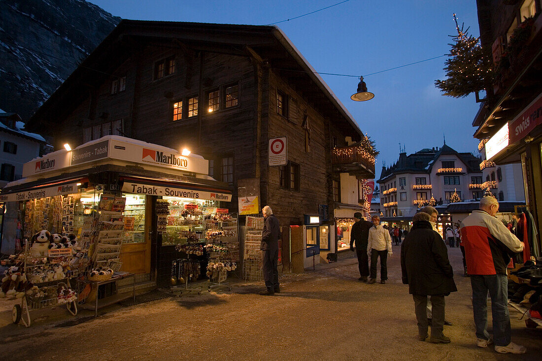 People walking over the illuminated shopping street Bahnhofstrasse in the evening, kiosk in foreground, Zermatt, Valais, Switzerland
