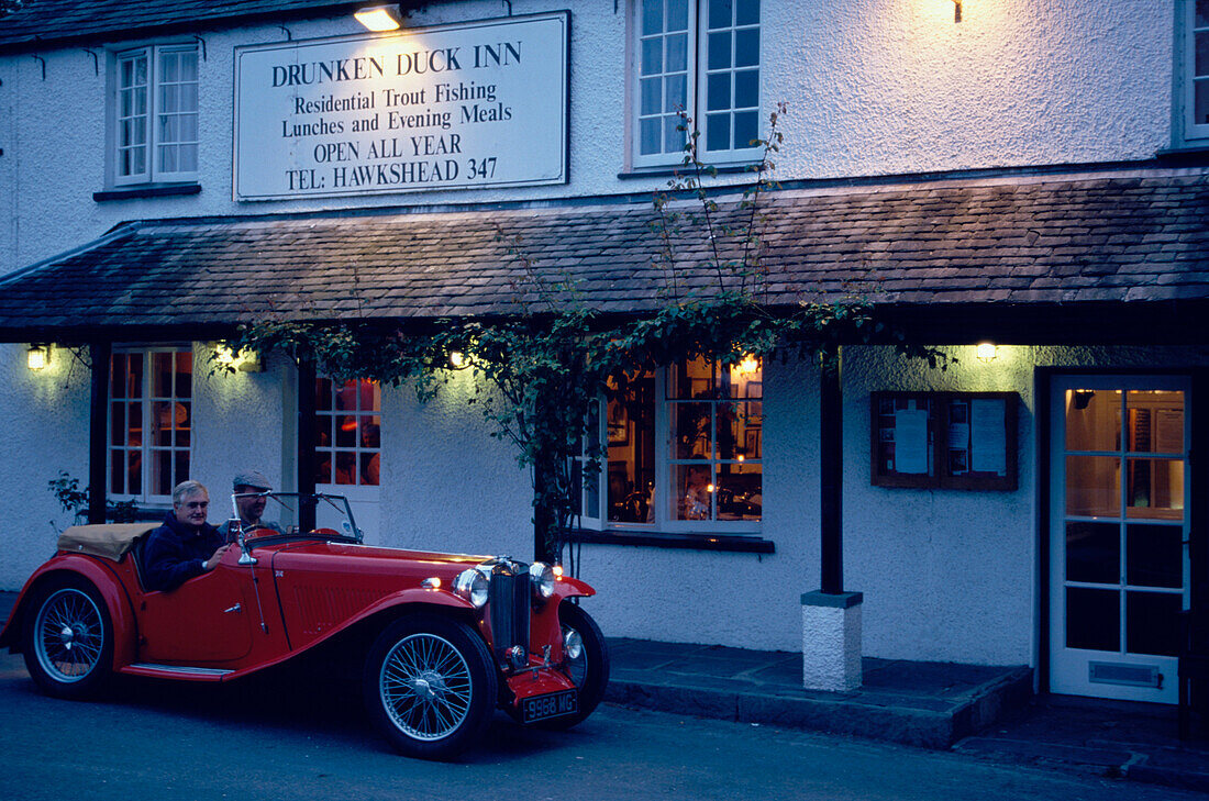 Two man driving an oldtimer in front of the Drunken Duck Inn, Lake District, Cumbria, England