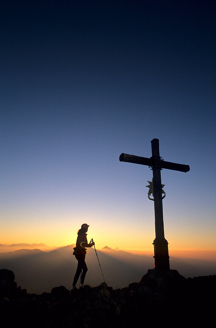 Bergsteigerin am Gipfelkreuz des Heubergs bei Sonnenuntergang mit Blick auf Wendelsteingruppe, Bayerische Voralpen, Oberbayern, Bayern, Deutschland