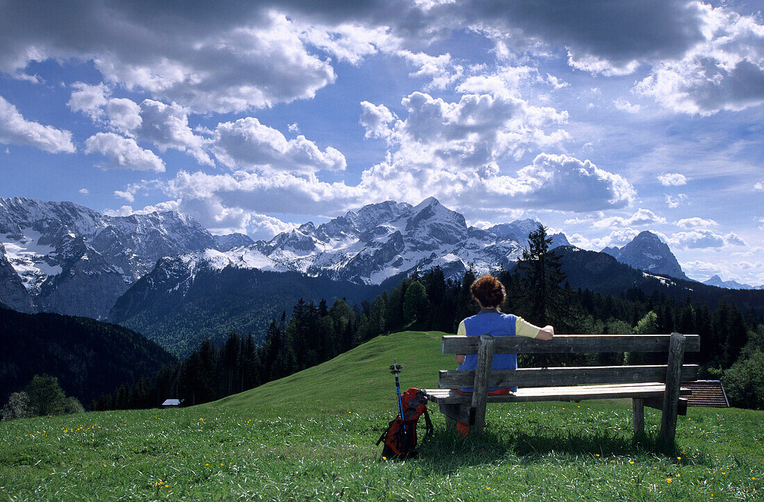 Hiker sitting on a bench at Eckbauer wih view to Wetterstein mountain range and Alpspitze, Werdenfels, Bavarian alps, Upper Bavaria, Bavaria, Germany