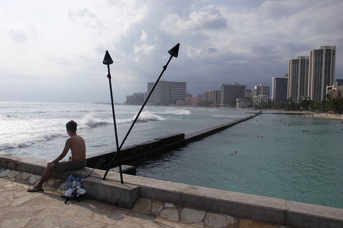 Boy sitting on a promenade with view at the sea and high rise buildings, Waikiki Beach, Honolulu, Hawaii, America, USA