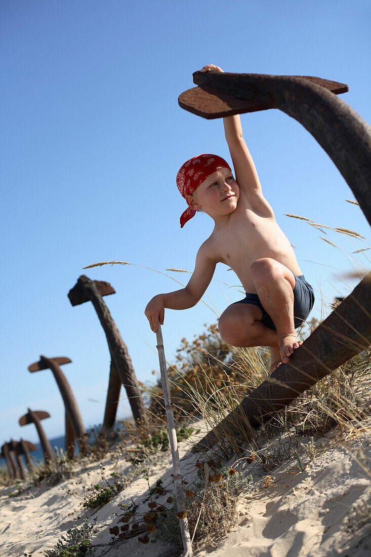 Junge spielt Pirat auf einem rostigen Anker am Strand, Ilha de Tavira, Tavira, Algarve, Portugal