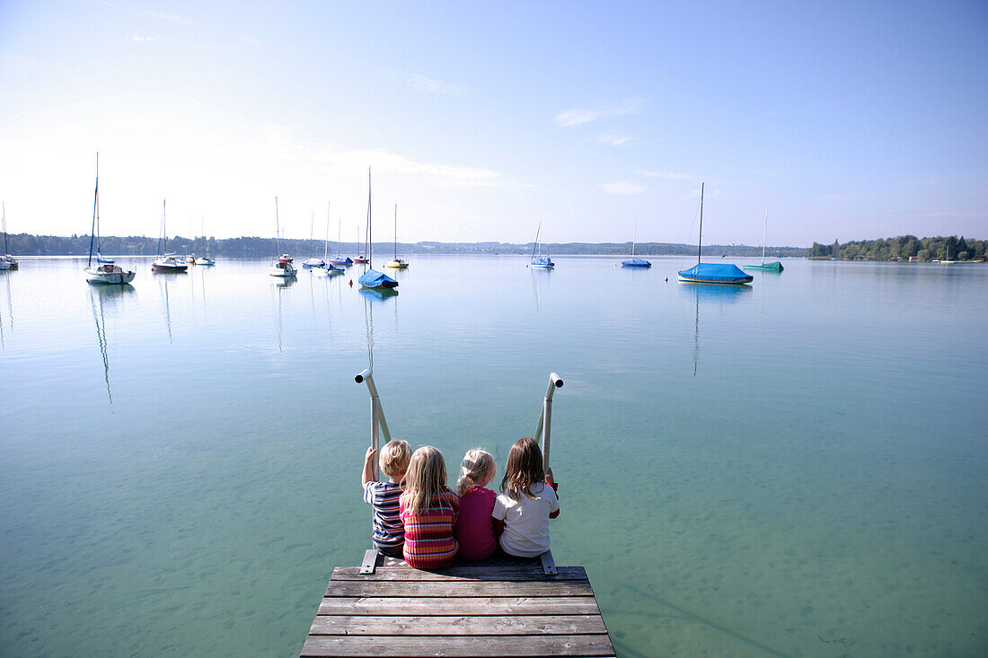 Vier Kinder sitzen am Rand eines Holzstegs, Walchstadt, Wörthsee, Bavaria, Germany