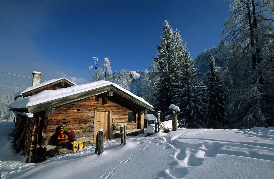 Wanderer mit Schneeschuhen bei Pause vor verschneiter Almhütte, Priesbergalm, Berchtesgadener Alpen, Oberbayern, Bayern, Deutschland