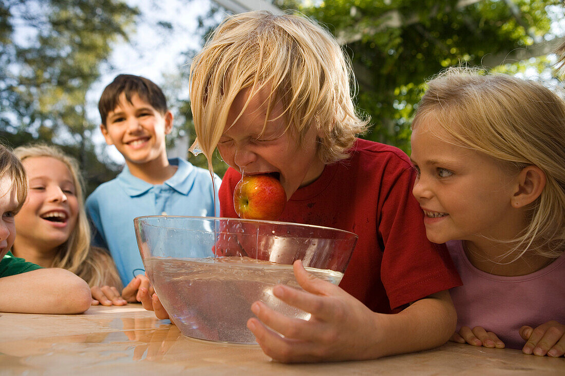 Junge mit nassen Haaren hält einen Apfel im Mund, Schüssel mit Wasser vor ihm, Kindergeburtstag