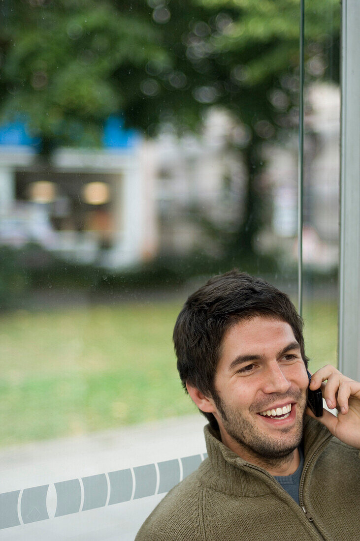 Junger Mann sitzt in einer Bushaltestelle und telefoniert mit einem Handy