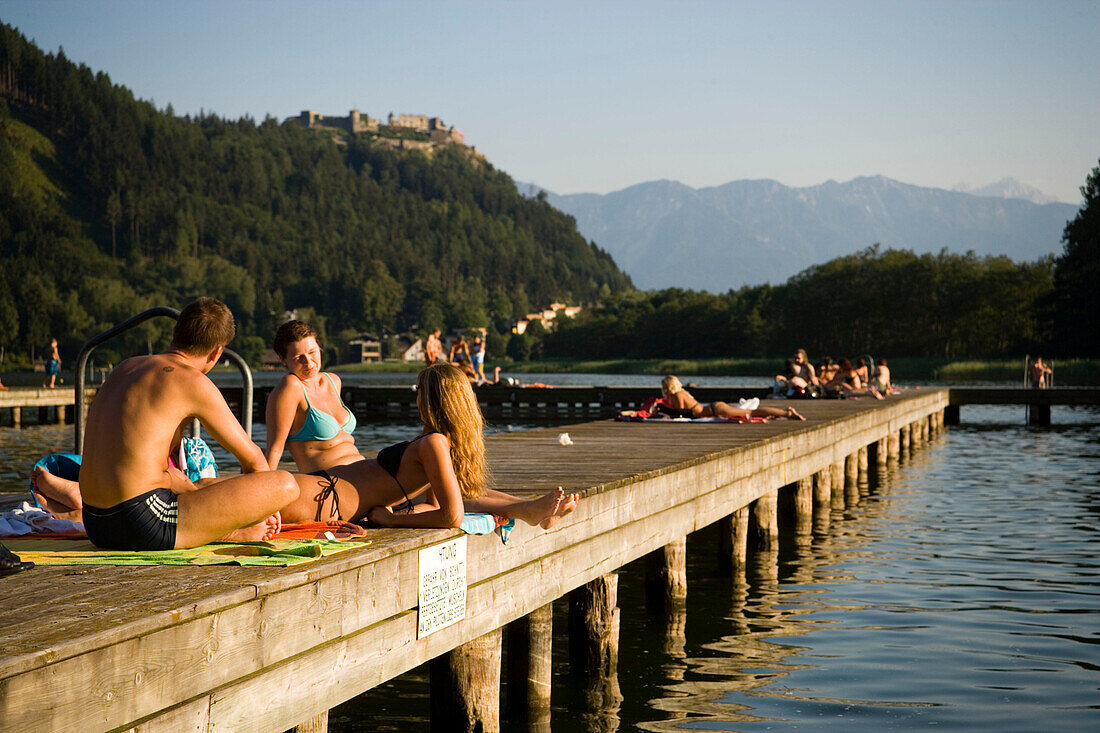 Young people sunbathing on boardwalk, Castle Landskron in background, Lake Ossiach, Ossiach, Carinthia, Austria