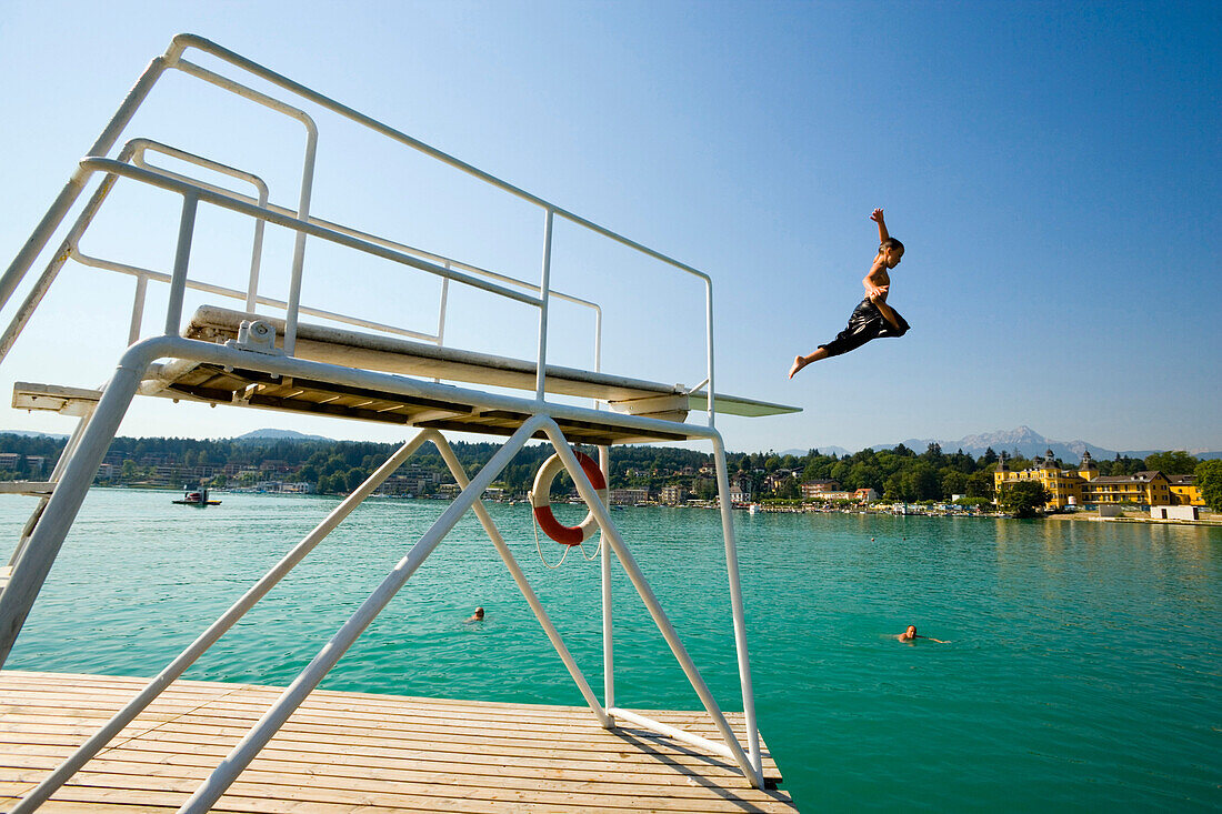 Boy jumping from diving platform into Worthersee, Hotel Schloss Velden in background, Velden, Carinthia, Austria