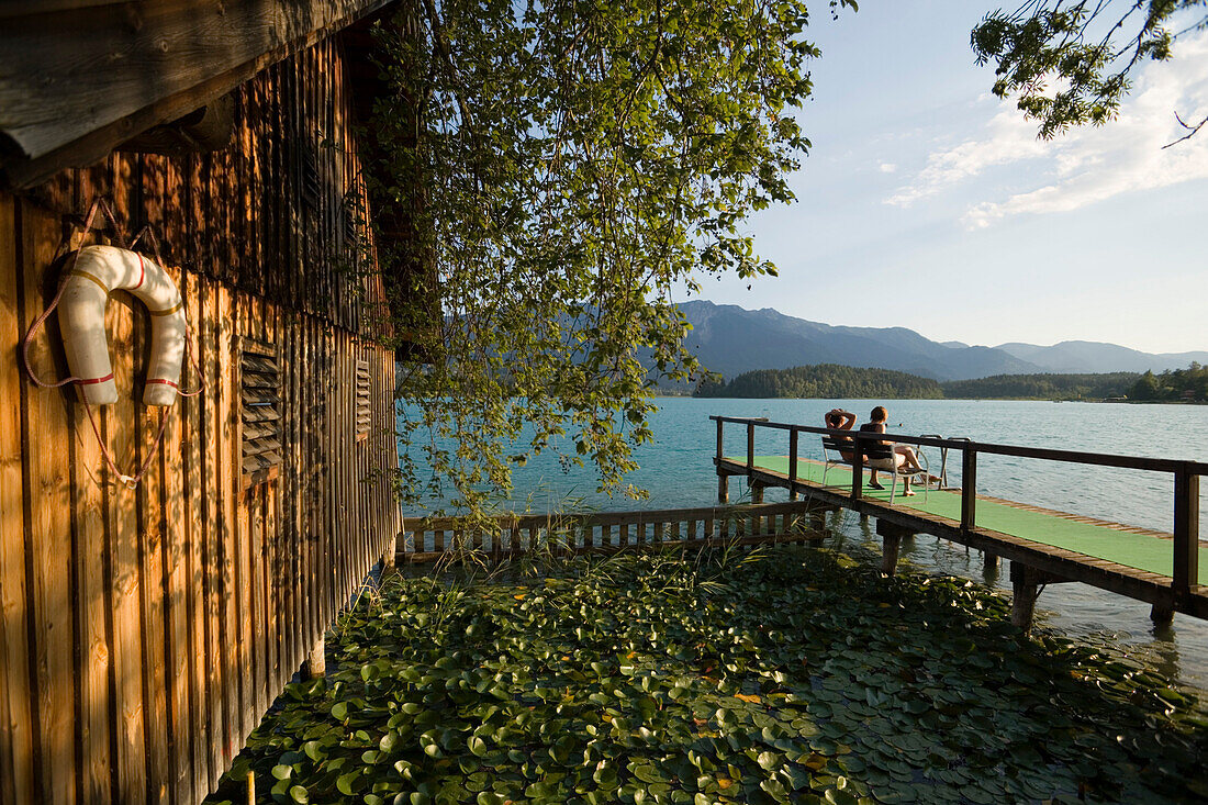Zwei Leute sitzen auf einem Bootssteg in der Nähe von einem Bootshaus, Faaker See, Kärnten, Österreich